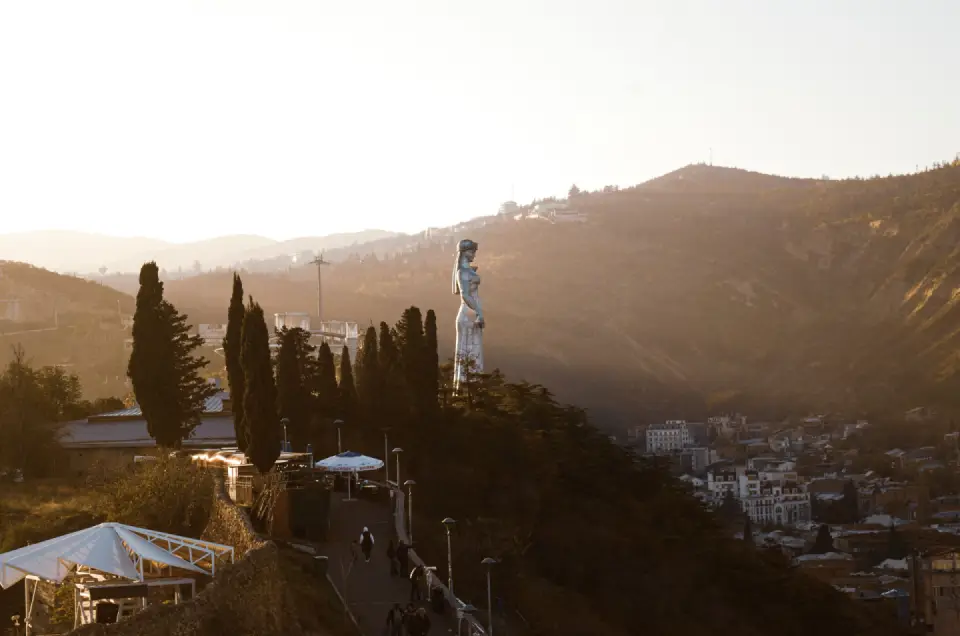 Hilltop monument surrounded by trees overlooking a Georgian city and distant mountains.