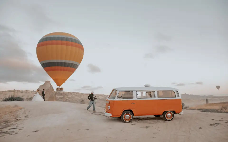 An orange vintage van parked in a scenic rocky landscape beneath a large hot air balloon, with a couple standing nearby.