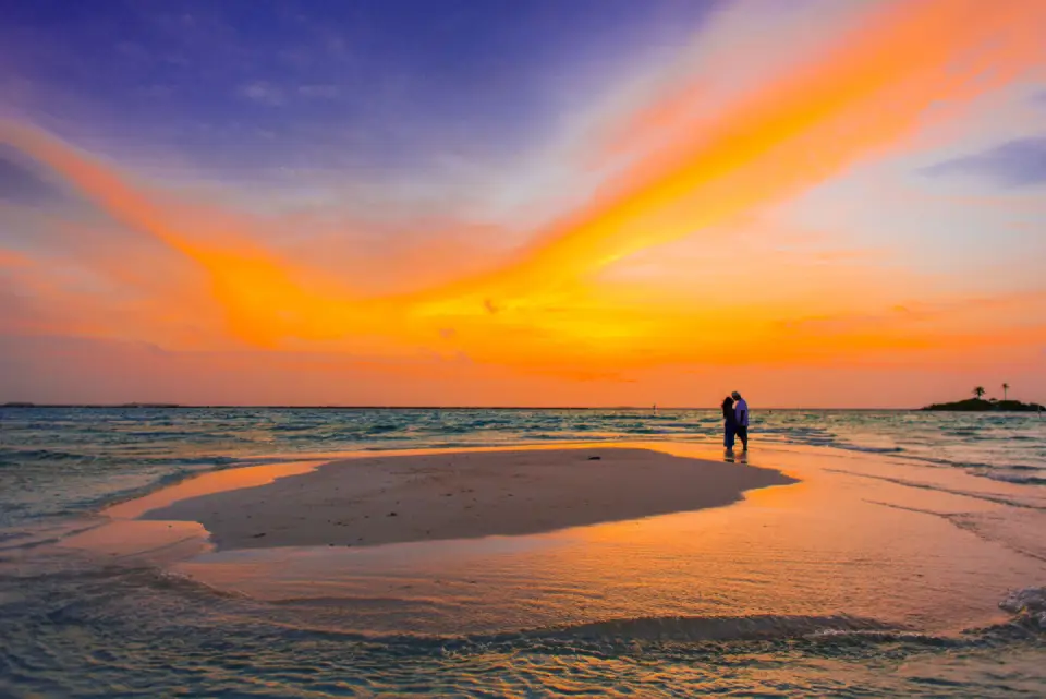 A couple standing on a sandbar surrounded by ocean water beneath a dramatic orange and purple sunset sky.