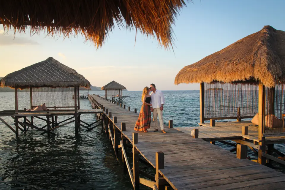 A couple sharing a romantic moment on a wooden pier between traditional thatched-roof gazebos overlooking the ocean at sunset.