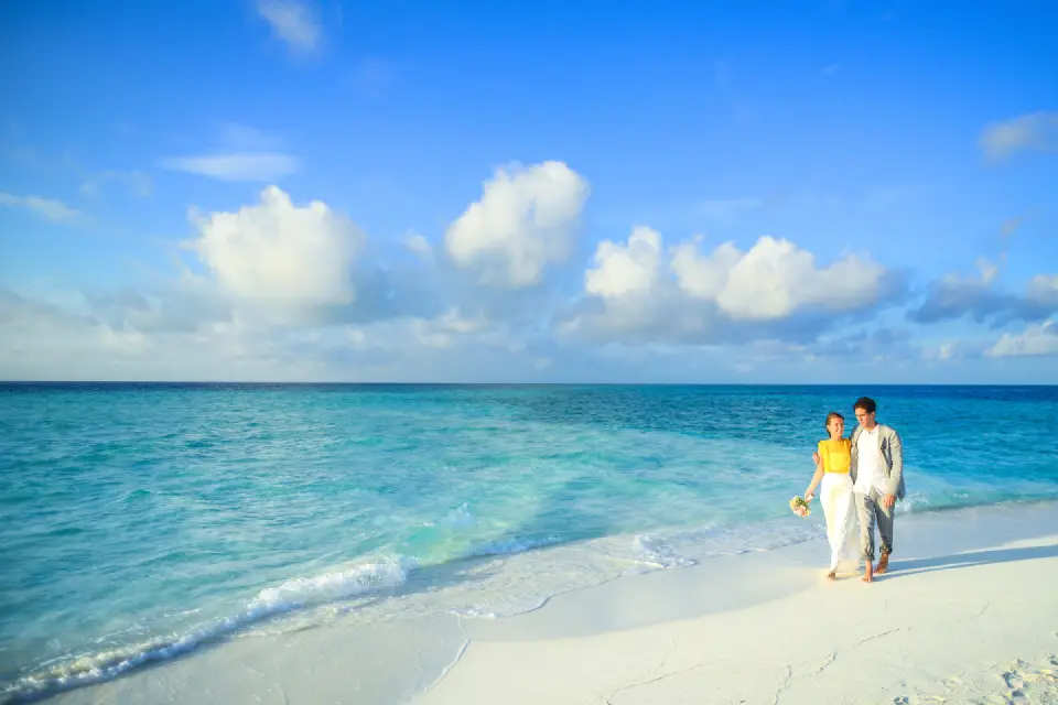 A couple walking barefoot along a pristine white sandy beach beside bright turquoise ocean water under a clear blue sky.