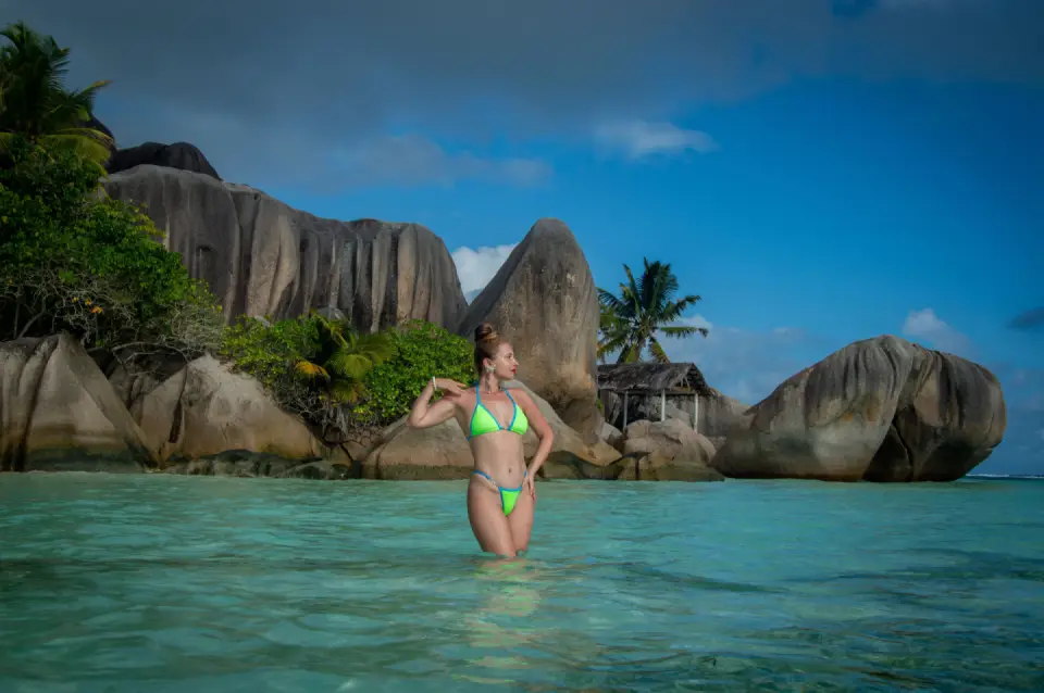 A traveller standing in a clear turquoise lagoon surrounded by giant granite boulders and lush green island scenery in Seychelles.