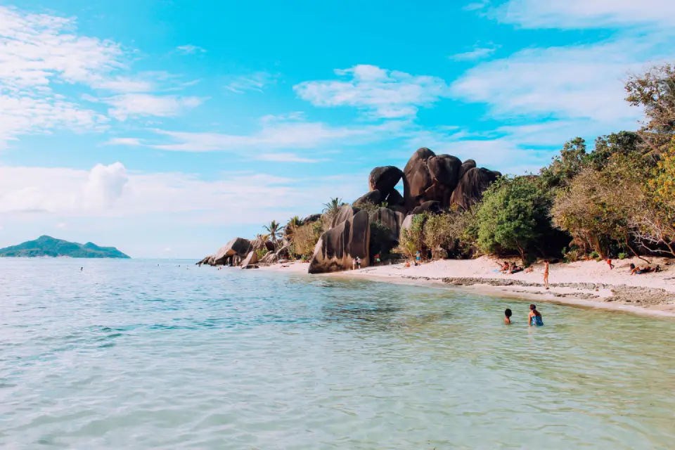White sandy beach with crystal-clear shallow water, granite boulders, and lush tropical vegetation in Seychelles.